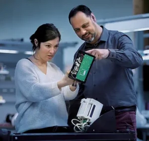 un homme à droite et une femme à gauche, debout, plan US, tenant un appareil électronique à mi-hauteur qu'ils regardent conjointement